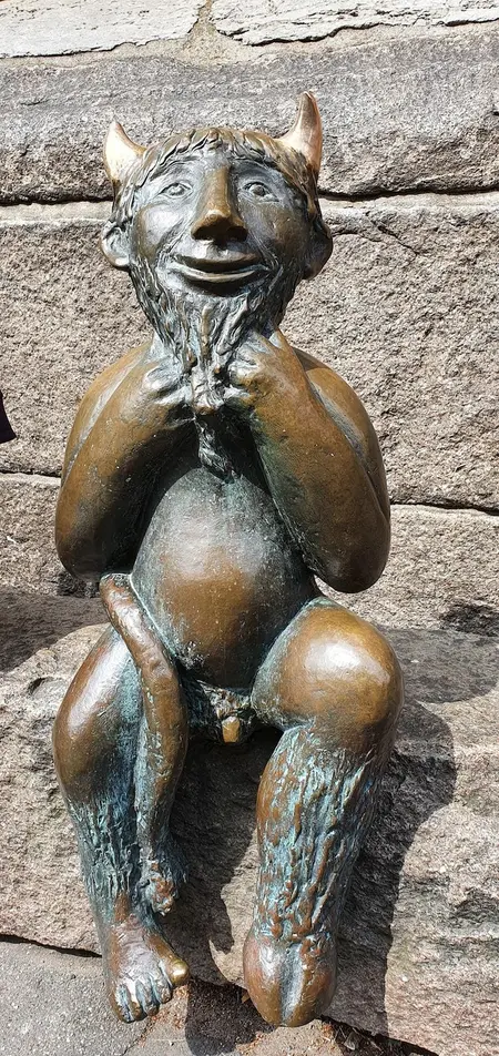 Bronzerne Skulptur des Teufels. Dieser sitzt auf einer Bordsteinkante und schaut den Betrachter mit menschlichem, freundlichen Antlitz an. Dennoch sind seine Hörner, der lange Schweif und eine Ziegenhufe deutlich sichtbar.